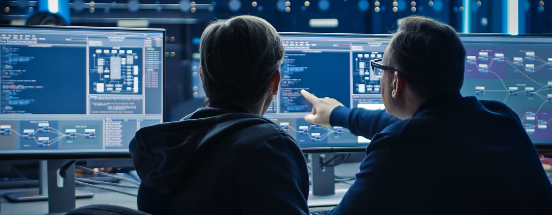Image shows the back of two men, one pointing at a computer screen with several computer screens in the background.