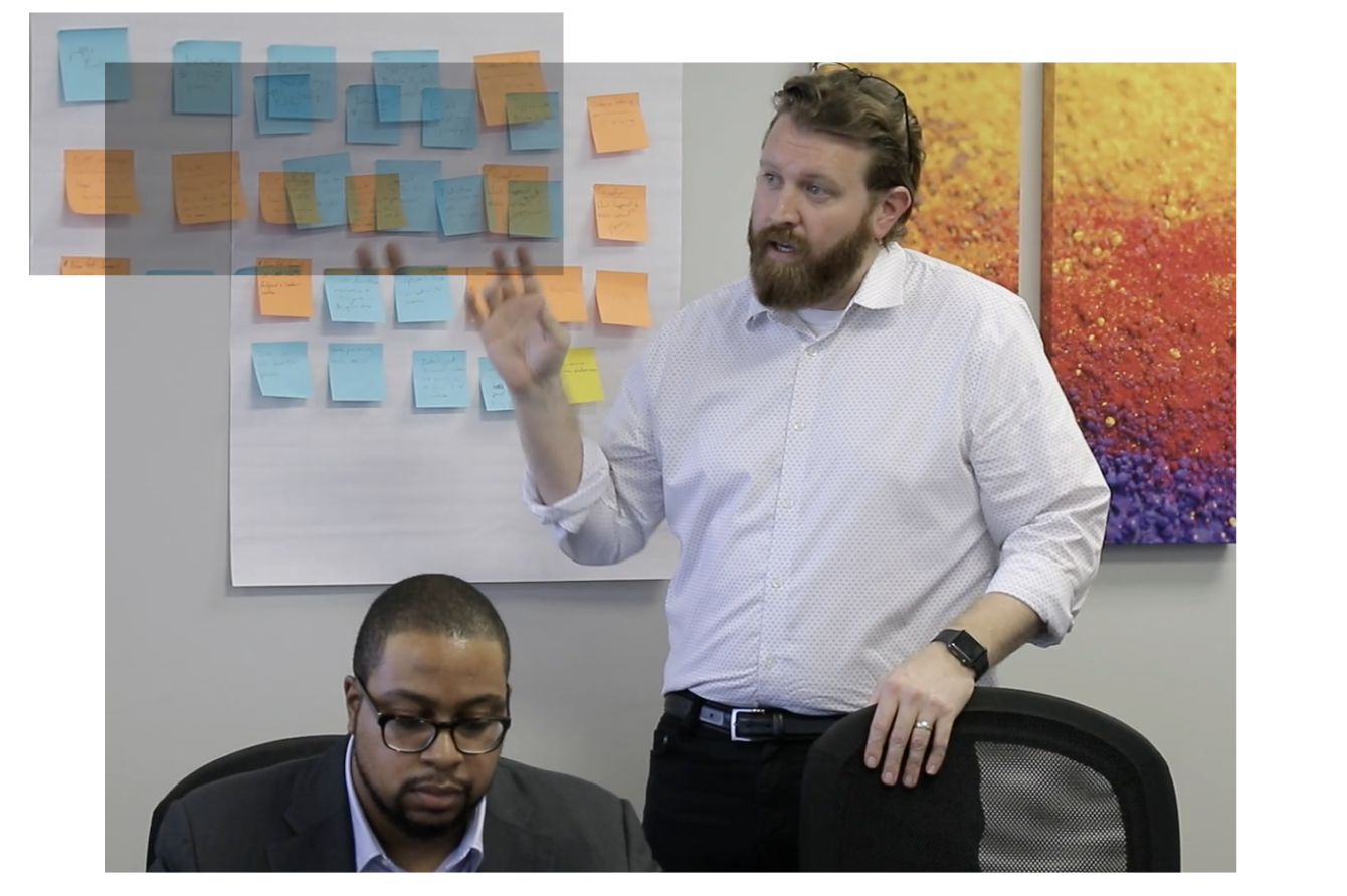 Image shows a man standing on the right and talking at a meeting with another man seated on the left. In the background, a wall is covered in Post-Its.