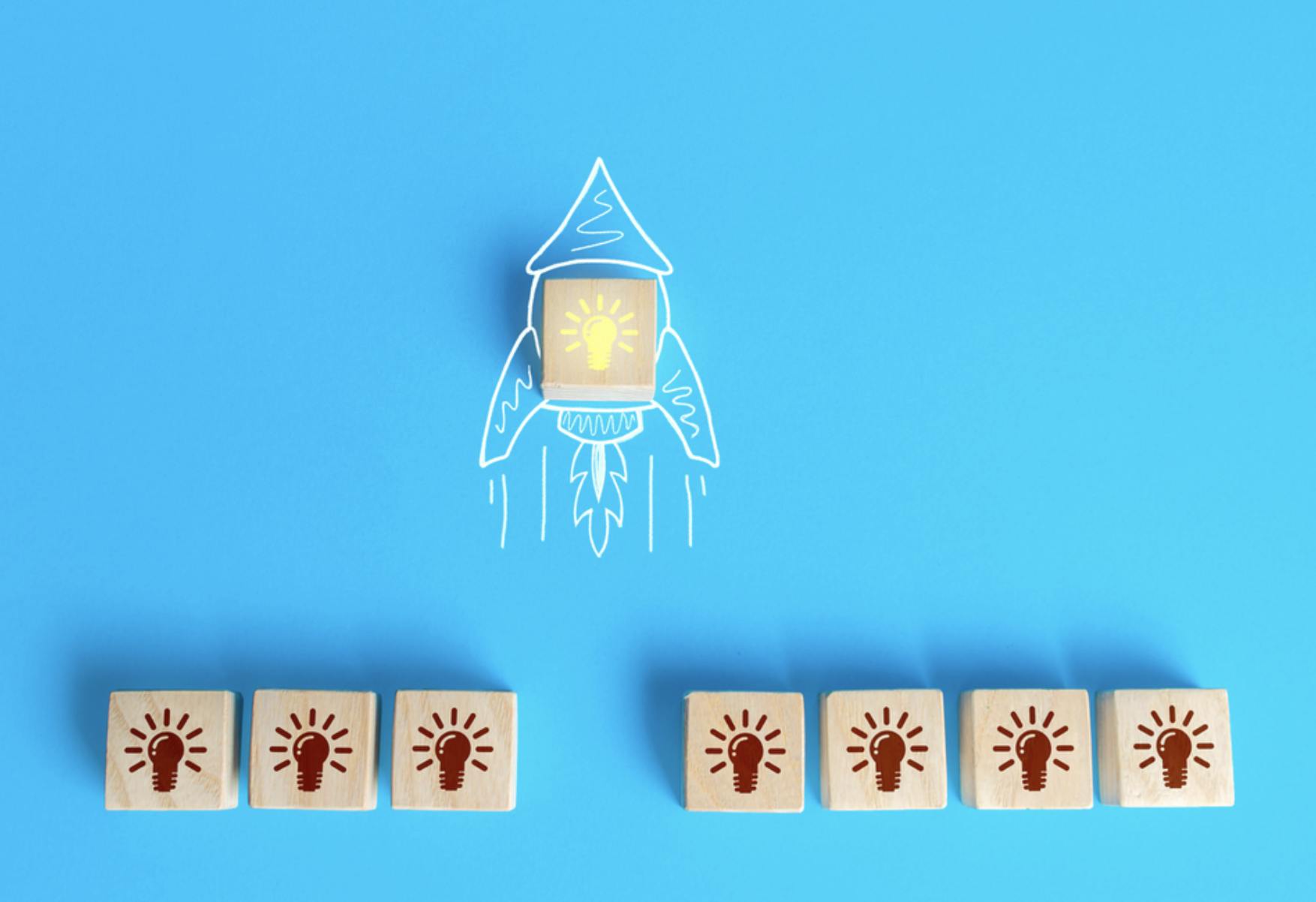 Stock Image showing a blue background with a rocket ship drawn in white. Seven wooden blocks all with a lit light bulb engraved in them in brown are lined up below the rocket ship with one block and in the center of the rocket ship drawing and the light bulb is painted yellow. Related to Pilot IRS project.