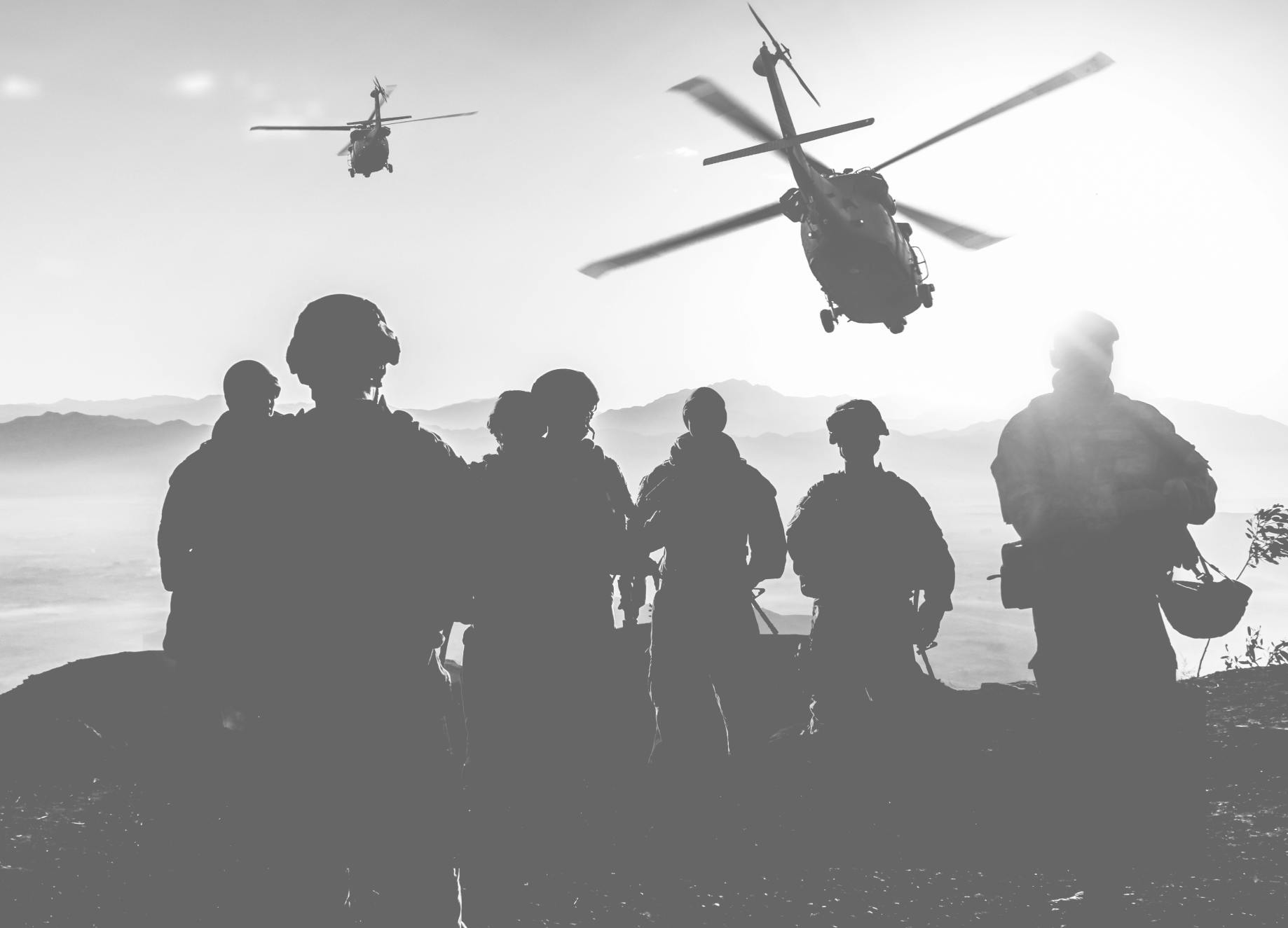Black and white image showing silhouettes of soldiers on top of a mountain with the sun shining brightly in the background and two helicopters flying towards the soldiers' position. Header image for military operations.