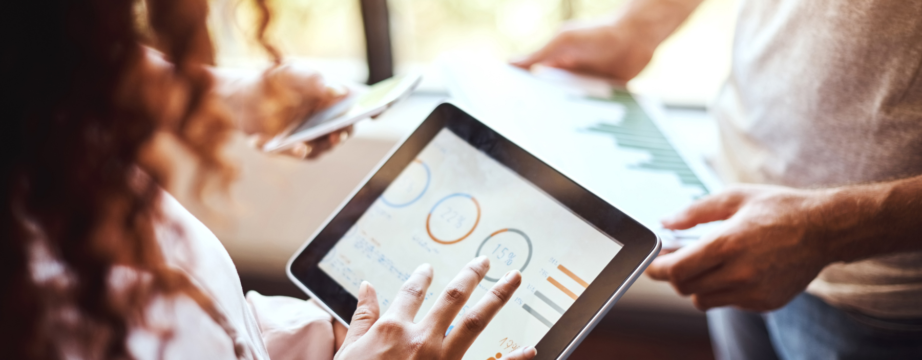 Image over looking a woman's shoulder as she parses through analytics on a tablet. Related to data services, government contractor, data services group, data recovery service, data visualization service, federal data services hub, data services network, and data services solutions.