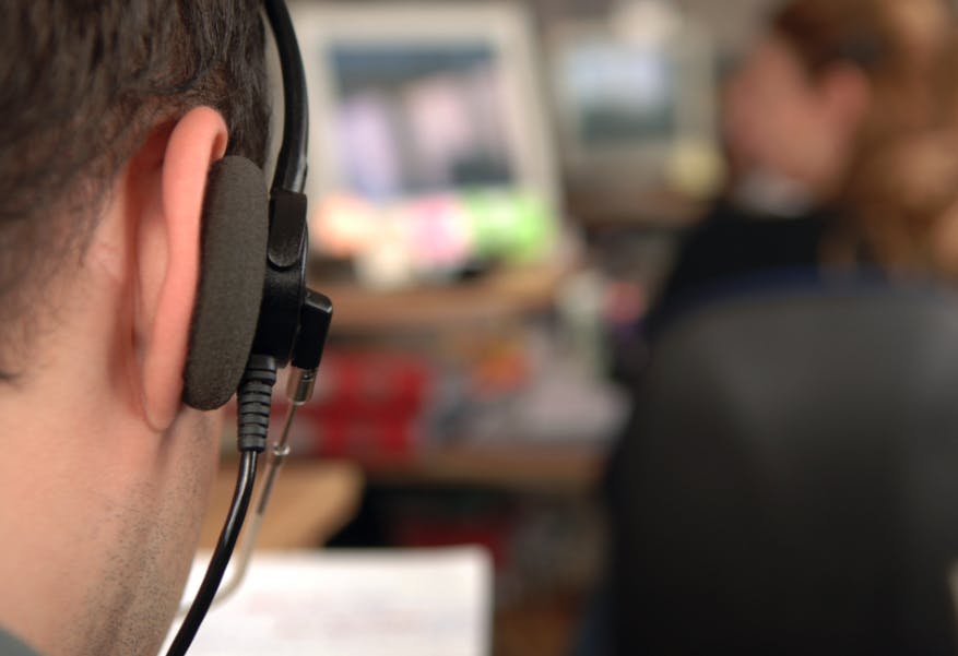 Image of the side of a man's head wearing a head set in the foreground and in focus. In the background, a woman sits at a desk and is out of focus.
