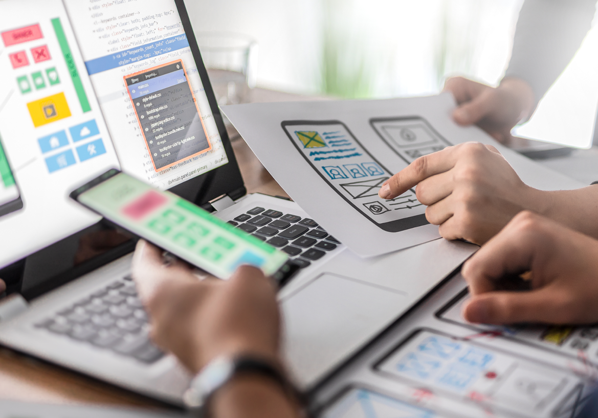 Image shows a person holding a cell phone above a computer with plans for an app on screen. Related to: app development, end user focused design, application development software, application development for cloud, application development federal.
