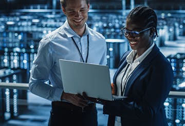 Image shows a man and a woman smiling together as they look at a laptop with server banks in the background. Related to: designing for future demand, architectural foundation, cyber architecture foundation, digital architecture services, digital architecture as a service,