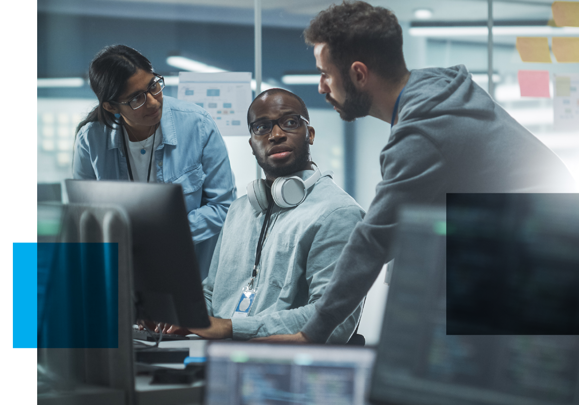 Image of a man sitting at computer station with headphones around his neck looking at another man beside him speaking with a woman in the group listening as well.