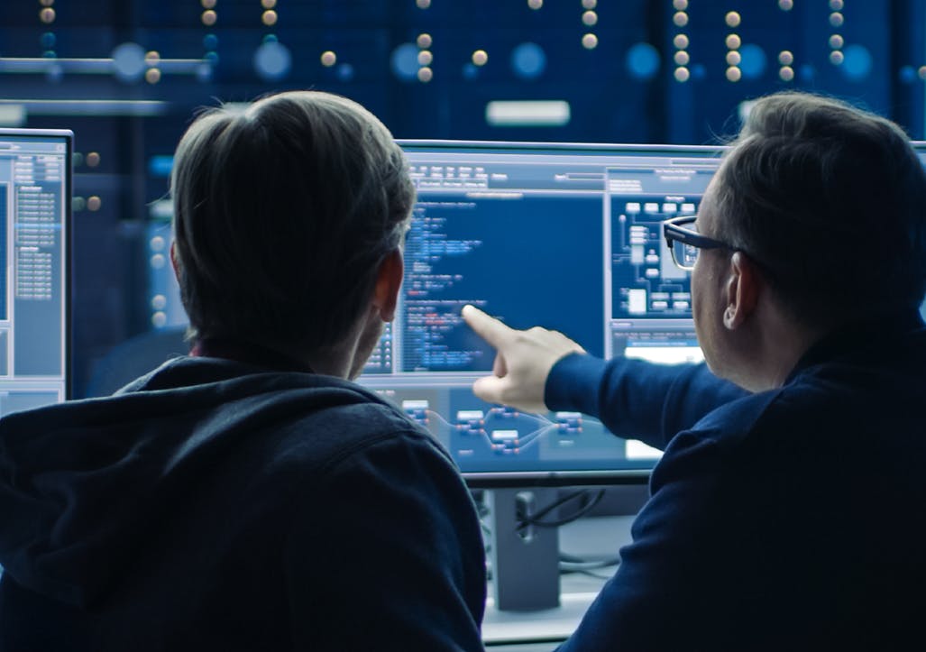 Image of two men with backs to the camera sitting at a computer screen, one pointing to code on screen. Related to cybersecurity, cyber operations warfare, security posture.