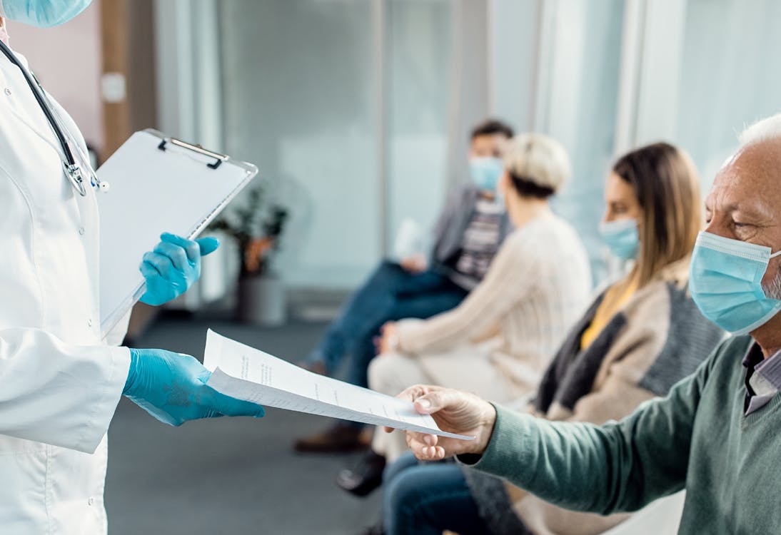Image of a waiting room with a doctor taking a piece of paper from an elderly man, while other patients wait in the background. Related to: corporate GWACs, federal cross-agency contracts, agency contract vehicles, SPARC, CTP BPA, NGITS Engineering BPA, CIT BPA, T4NG IDIQ, VICCS IDIQ.