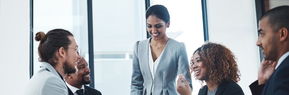 Image of an Indian woman standing in a corporate setting surrounded by colleagues.