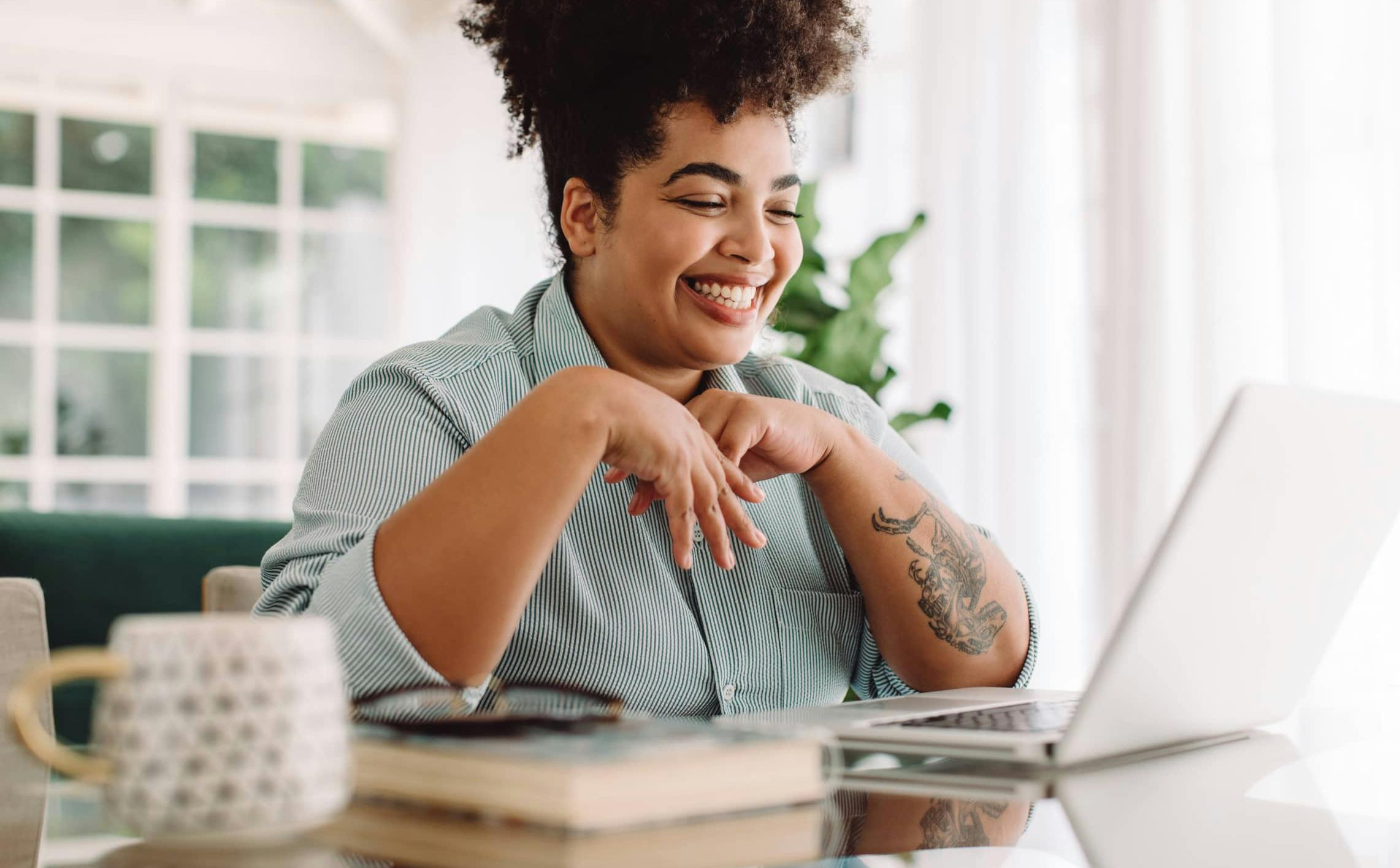 Image of a smiling woman with a tattoo on her forearm gazing down at a laptop in her home.