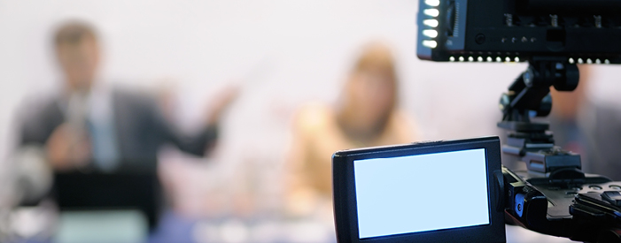 Image shows in the foreground a camera and screen in focus while in the background three people sit out of focus.