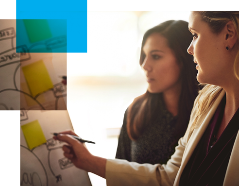 Image shows two women conferring over a white board with a plan on it.
