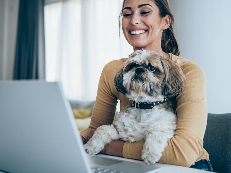 Image of a woman with her fuzzy dog on her lap, smiling and looking at her computer screen in a house setting. Related to: GovCIO Pets life, pets at work, pets culture, pets appreciation