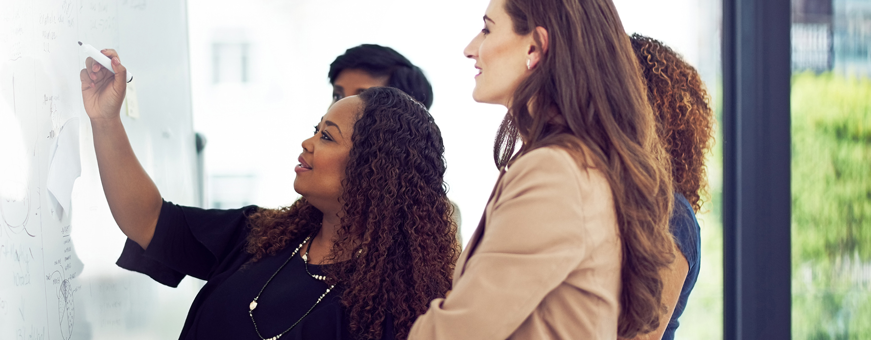 A woman stands at a white board poised to write with three team members standing by watching. Related to: effective and optimized operations, innovative operating models, strategic partners, Agile implementation, agile integration, agile supply chain, DevSecOps framework, operational best practices, future-ready organization.