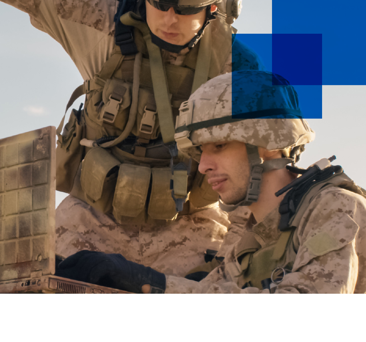 Two male soldiers in desert camouflage uniforms lean over a lap top while looking at the screen.