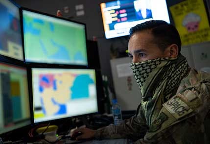 Image of a soldier with his mouth covered by a handkerchief looking at multiple computer screens. Related to: Kessel Run, software factories, Kessel Run software factory, DOD software factories, Devsecops software factory