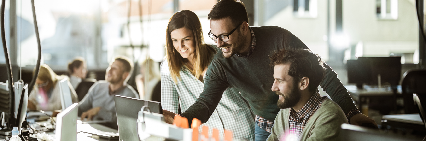Image of two men and a woman looking at a computer screen in an office. One man is standing and pointing at the screen while everyone smiles.
