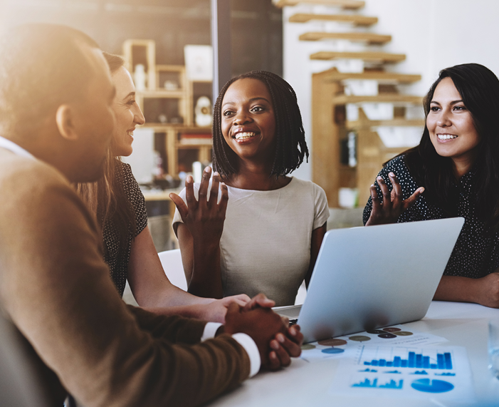 Image of a group of people at a table with a Black woman in the center, sitting in front of an open laptop and talking. Two more women and a man around her sit, listening. Related to: social responsibility, corporate responsibility, volunteering, corporate community engagement