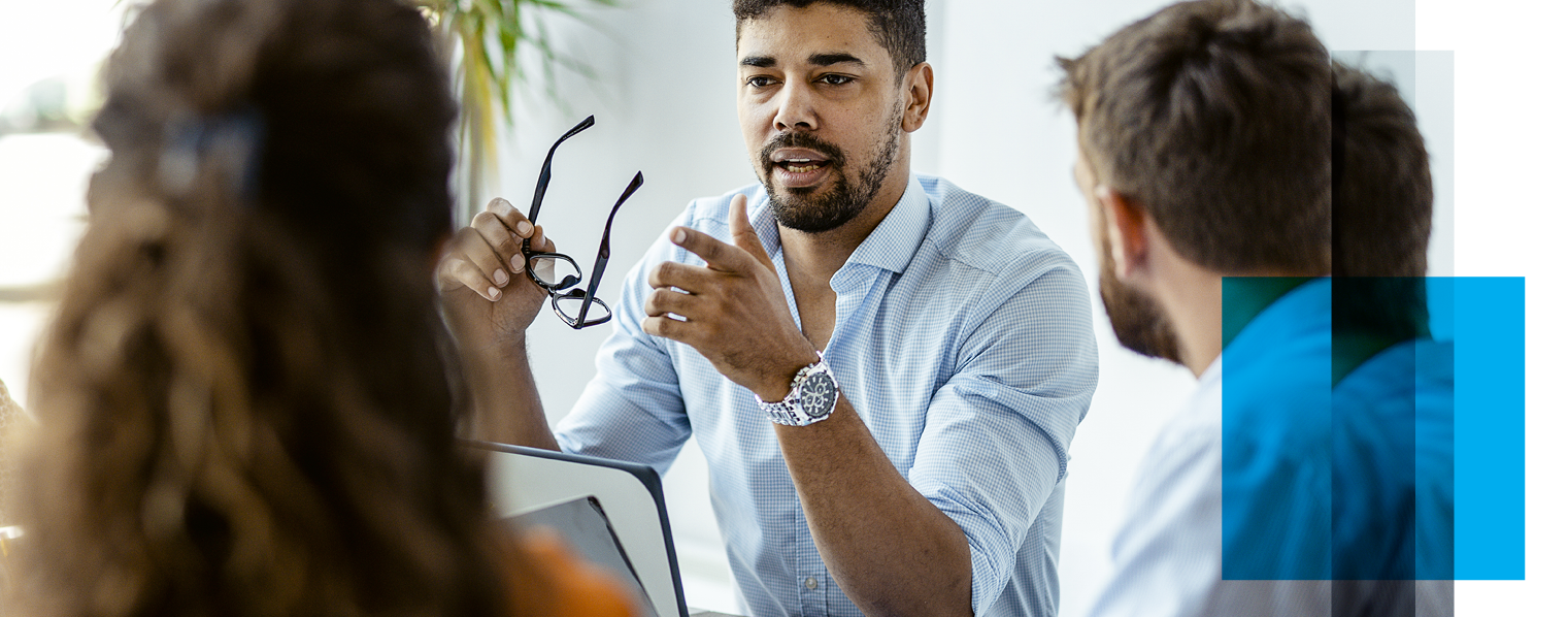 Image shows a man facing the camera, holding eye glasses and speaking with another man and a woman with their backs to the camera listening to the speaker. RElated to: our work, GovCIO statistics, company culture, company makeup, company demographics, company status