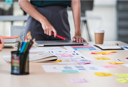 Image of a woman standing at a table with post it notes and documents organized on top, pointing at one. Related to: agile transformation, transparency culture, agile trainers, learning organization, continuous incremental growth.