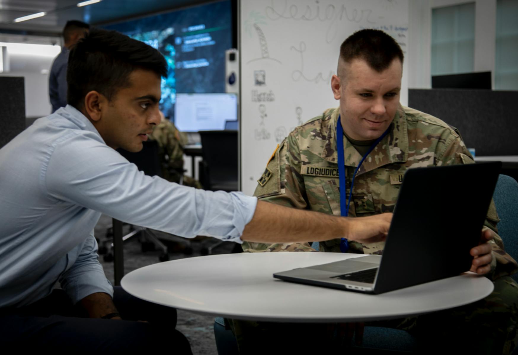 Image shows two men sitting at a table looking at a laptop, one pointing to the screen, the other in fatigues. Related to: software factories, software factory, dod modernization, digital transformation, rapid data sharing, secure data transfer, defense space, army software factory, cohort 6.