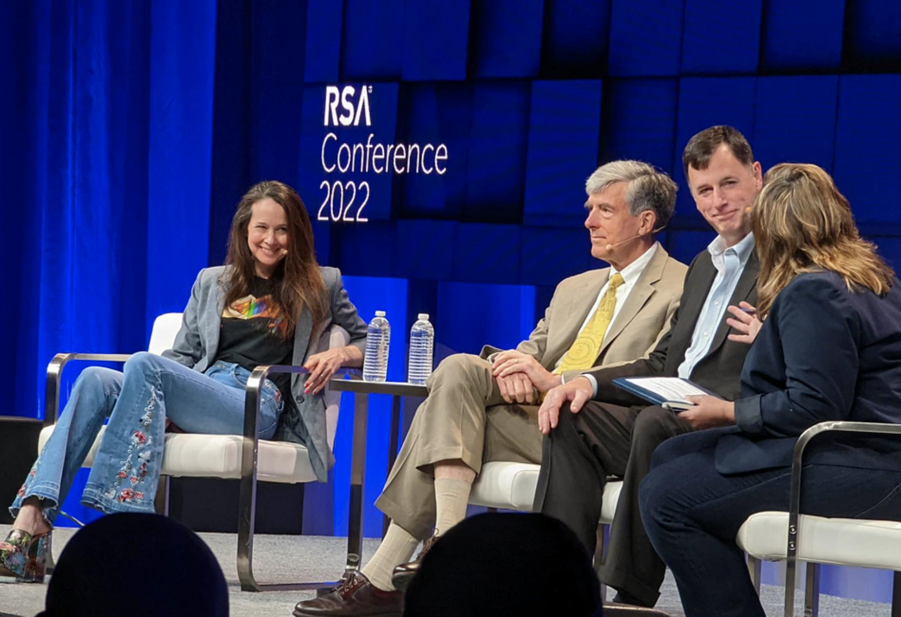 Image of the stage at RSA Conference 2022 with two women and two men sitting on a panel. Related to cyber defense, CISA Shield Up effort, national security defense, cyber security engineer, cyber security jobs, cyber security definition, cyber security framework, and cyber security threats.