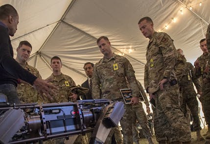 Image of soldiers in camouflage uniforms in a tent standing around a table listening to another man not in uniform at the table who is speaking.