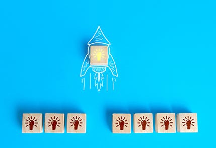 Stock image with a blue background, a rocket ship drawn in white and a wooden block with a yellow light bulb in the middle of the rocket ship. Seven other wooden blocks with brown light bulbs are lined up at the bottom of the image.