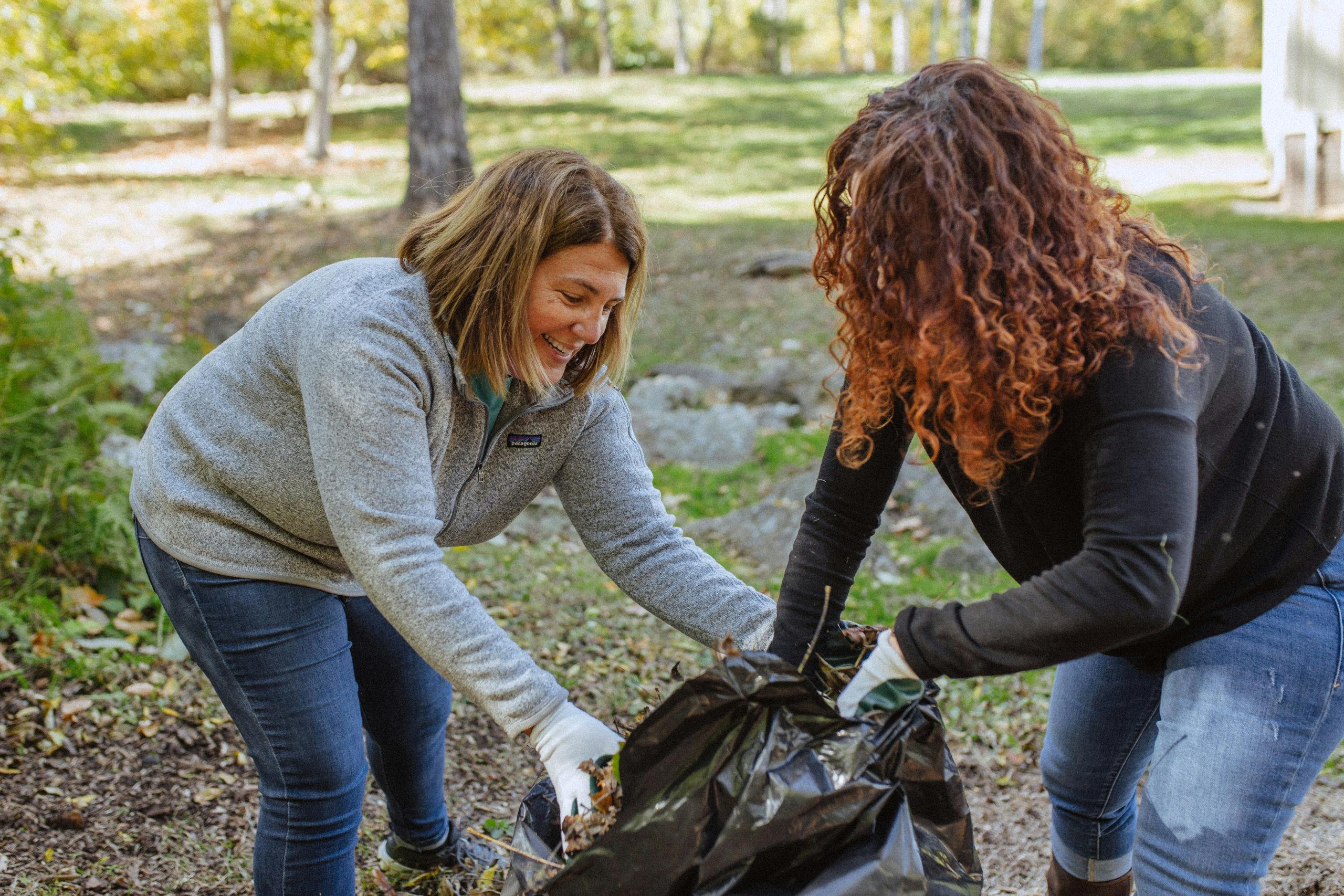 Image of two women gathering fall leaves in a trash bag together.