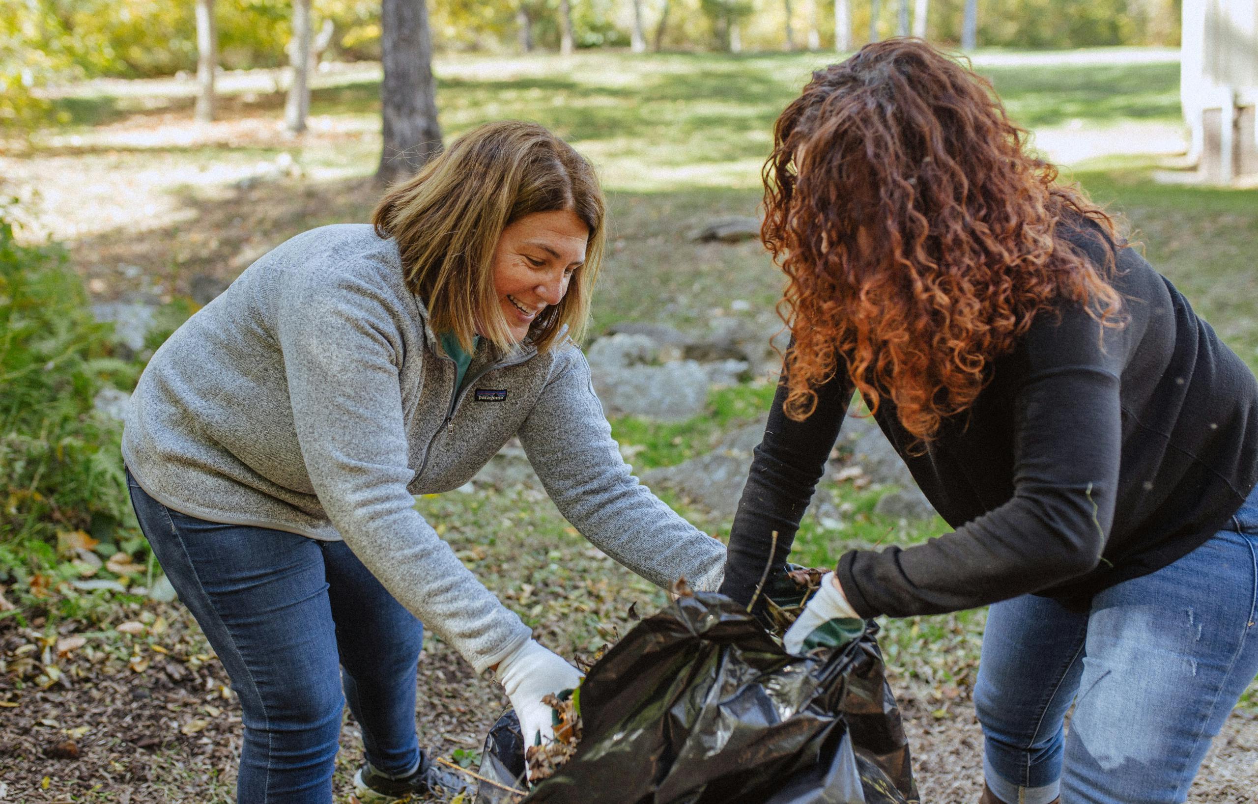 Image of two women gathering fall leaves in a trash bag together.