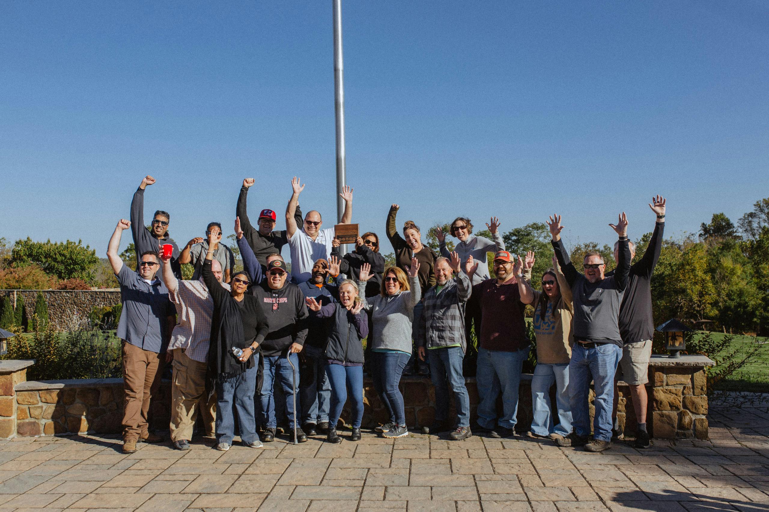 Image of a group of people raising hands as they look at the camera. Related to: vets giving back, hiring vets, hiring veterans, government contractor hiring veterans, charity work, charity work for vets, vets supporting vets