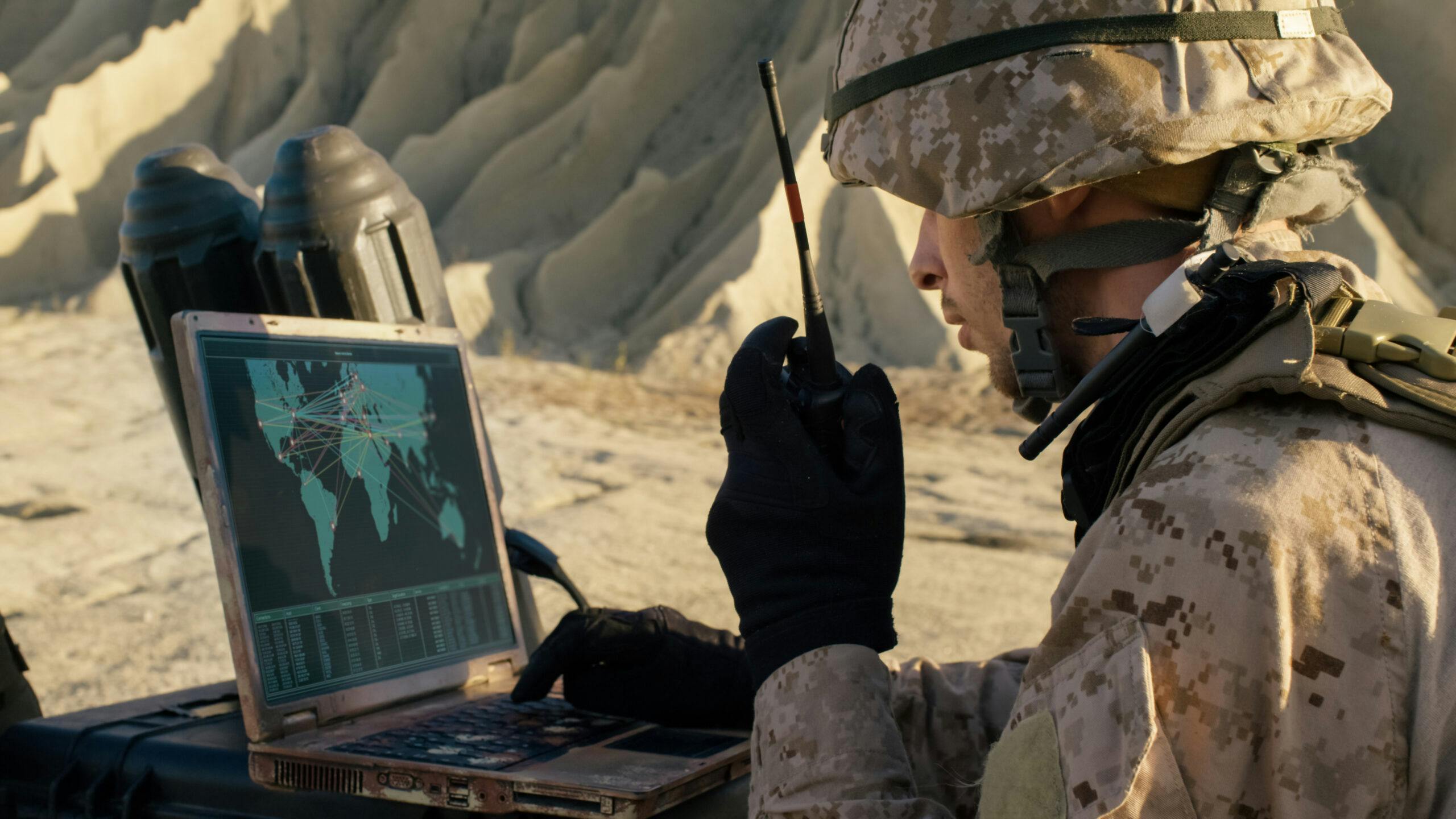 Image of a soldier talking into a walkie-talkie while looking at a laptop screen with a global map on it.