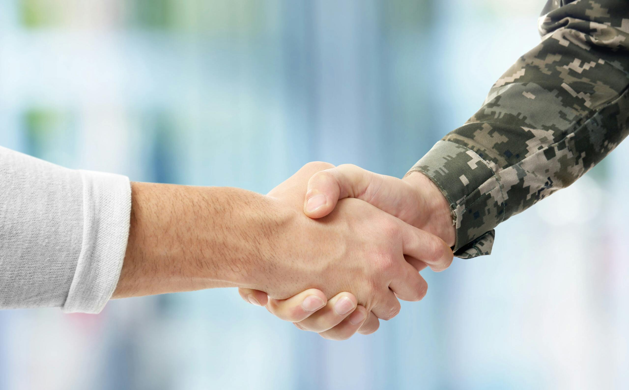 Image of people shaking hands. One arm is covered in army fatigues, the other has a white shirt on.