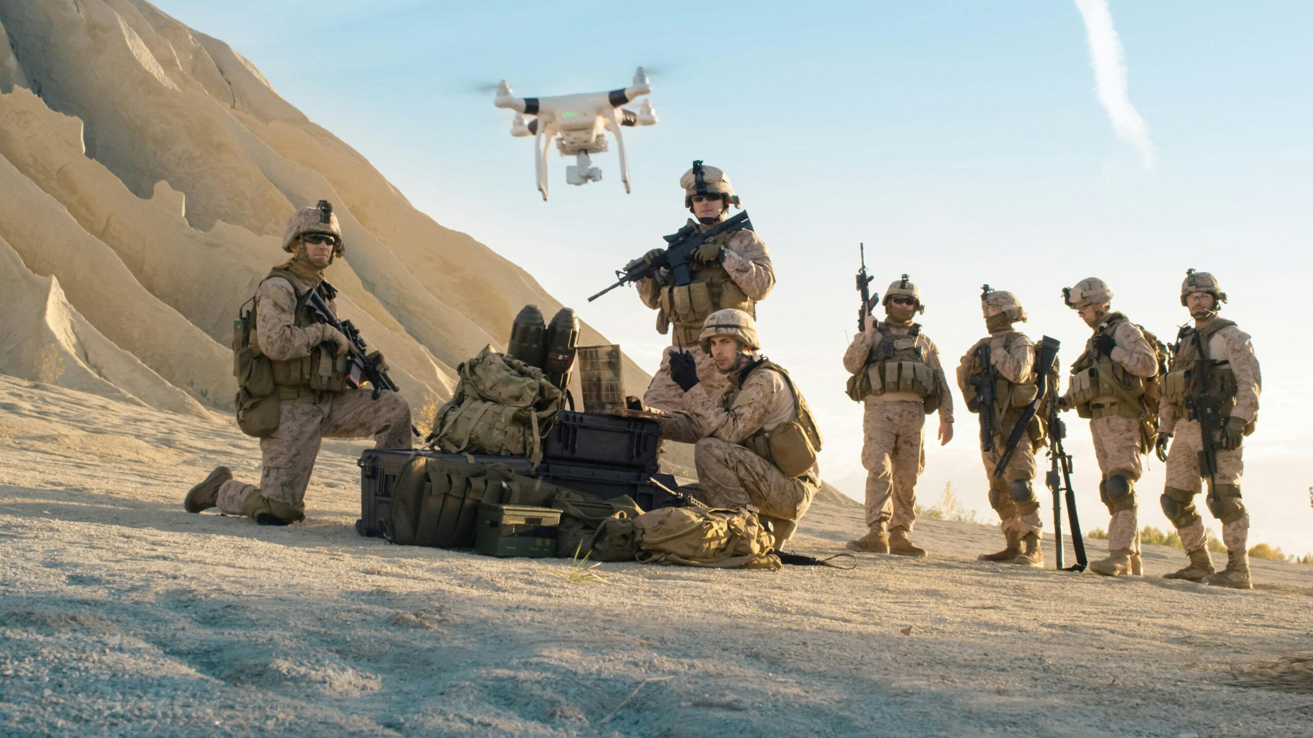 Image of soldiers in full tactical gear in the desert with a dron hovering overhead, gathered around a field laptop.