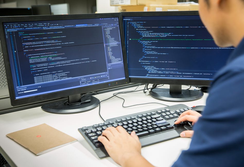 Image looking over shoulder of a person at two computer screens with code up. Related to: federal open-source software, open-source software in government, open-source software defense space, devsecops and open source, open source tech.